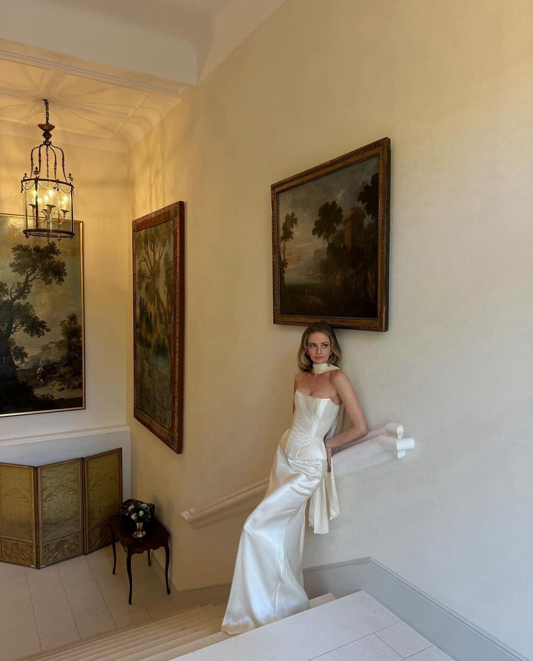 a woman in a white wedding dress stands on a staircase, leaning on the railing. the dress is a satin corset top with a flowing skirt and a white scarf around her neck. she is looking to the side. behind her are two paintings hanging on the wall.