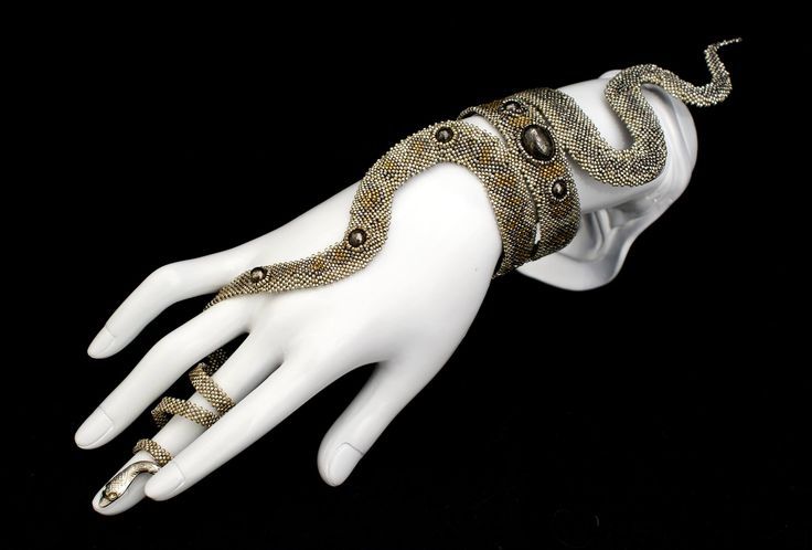 a snakeshaped beaded bracelet and ring set is displayed on a white mannequin hand against a black background. the bracelet wraps around the wrist and extends up the arm, resembling a snake. the ring is also snakeshaped and coils around the finger. the jewelry is made of gold, silver, and brown beads. the ring has a snake head with white eyes.