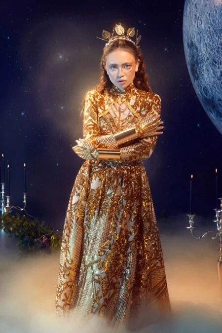 a woman with a gold crown is wearing a glittering gold dress standing against a backdrop of  a moon and stars. she has on golden bracelets and gloves.  she looks like a queen,  a  goddess, or a  fairy.  the background is a night sky, filled with stars and a full moon. there are candles and a candelabra nearby.