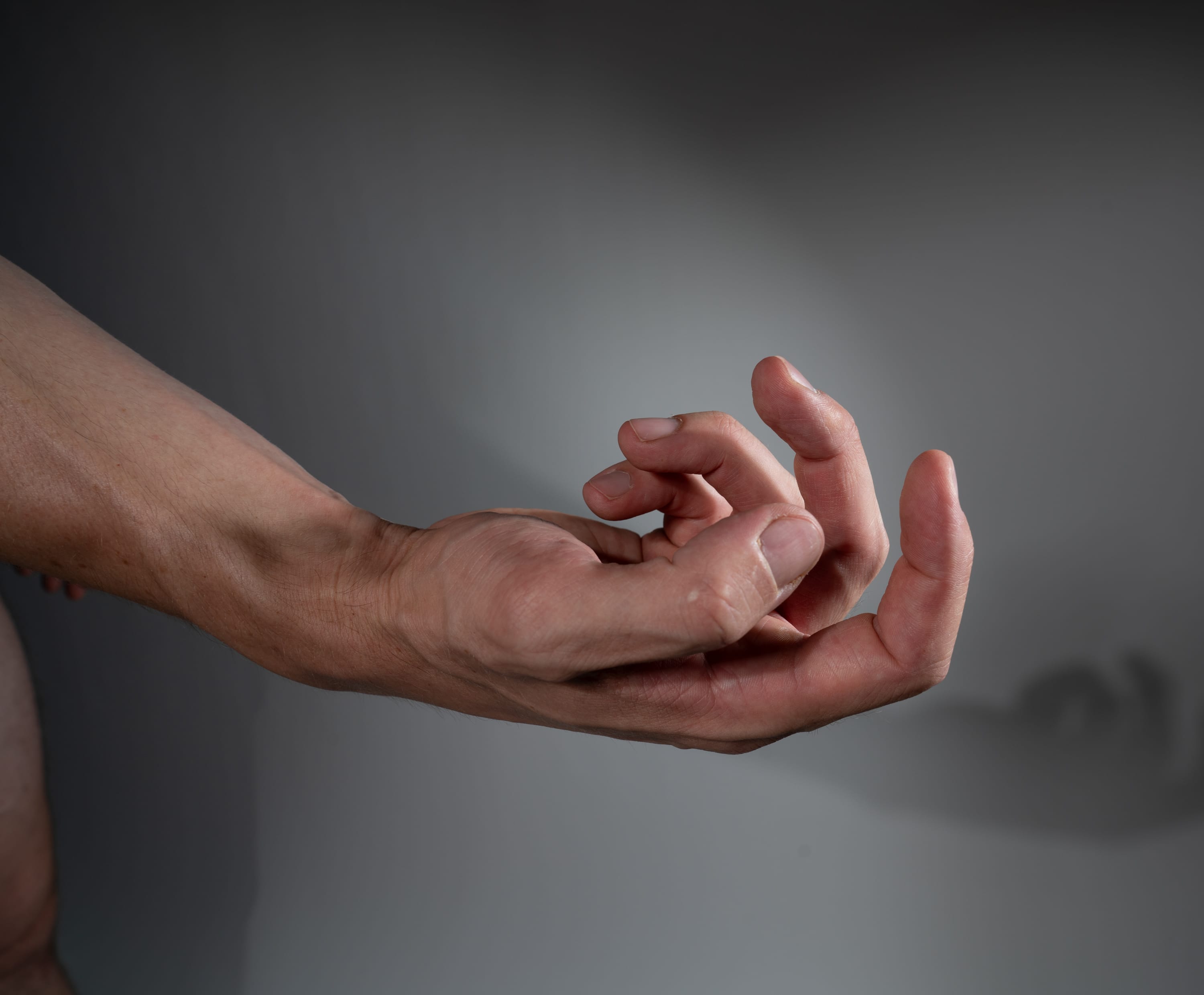 the image shows a man's hand and arm extended against a gray background. the hand is partially clenched, with the fingers curled inward. the lighting is artificial, creating a spotlight effect on the hand and casting a shadow on the background.