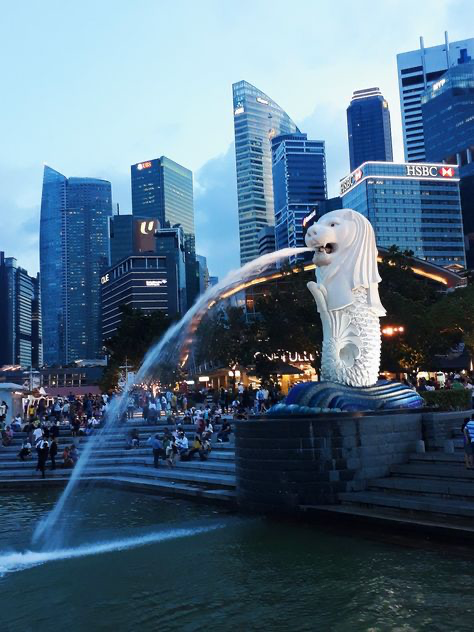the image shows the merlion statue in singapore at dusk. the iconic merlion fountain is in the foreground, with water spraying from its mouth.  behind it, the modern singapore skyline is visible, with numerous tall skyscrapers illuminated against the darkening sky.  the scene includes a crowd of people gathered in the plaza around the fountain. the hsbc building is prominently featured in the skyline.  the overall mood is serene and captures the beauty of singapore's cityscape at twilight.
