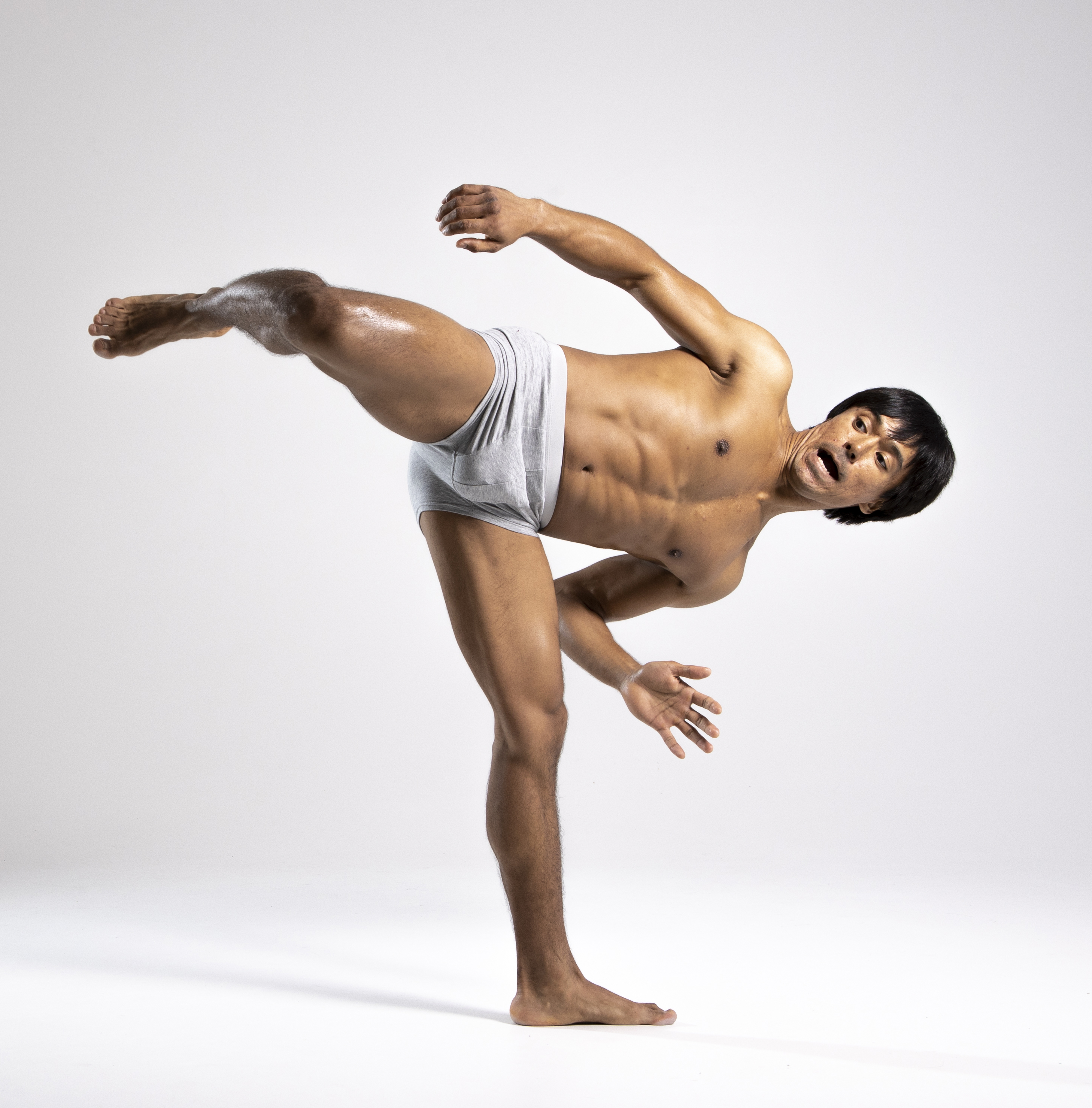 a shirtless man in gray briefs is captured in a dynamic pose against a plain white background. he's balancing on one leg with the other extended high, arms outstretched, his face expressing intensity. the image conveys a sense of athleticism, flexibility, and the art of contemporary dance.