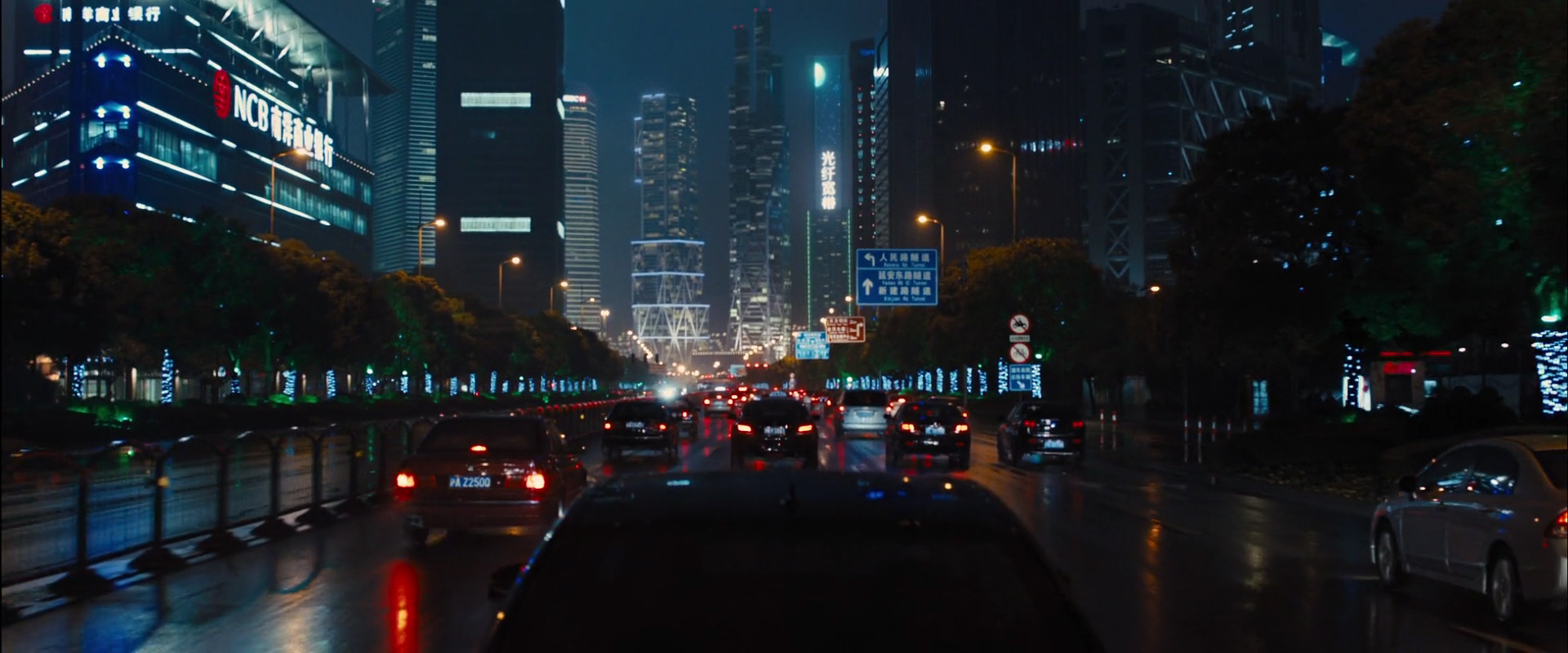 a wide shot of a busy city street at night, with a long line of cars driving down the street. the street is wet from rain and the buildings are all lit up in the background. there are tall skyscrapers in the background, creating a dramatic cityscape.  the image is taken from the backseat of a car. the focus is on the car in front.
