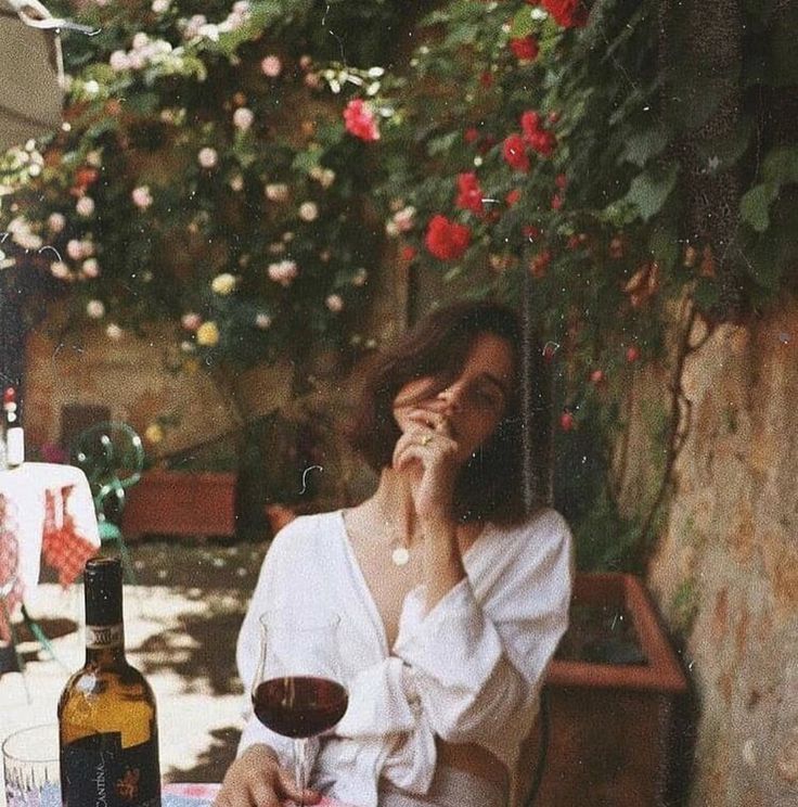 the image depicts a young woman with short brown hair, sitting outdoors in a lush garden filled with red roses. she's wearing a white, loosefitting shirt and a simple necklace.  she's holding a glass of red wine, appearing relaxed and pensive. the setting is serene and romantic, with dappled sunlight and shadows creating a warm, vintage aesthetic. the overall mood is one of quiet contemplation and peaceful enjoyment. the image has a slightly grainy, filmlike quality that enhances its vintage feel.