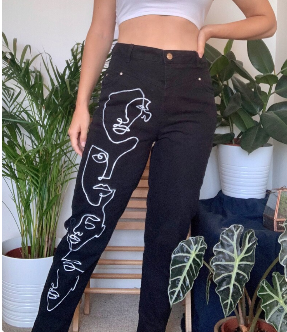 a person is modeling a pair of black jeans with white line art faces drawn down the left leg. the jeans are highwaisted and the model is wearing a white crop top. there are several green houseplants visible in the background.
