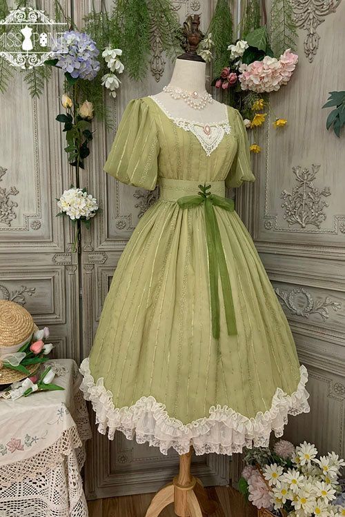 a green vintagestyle lolita dress is displayed on a mannequin. the dress features puff sleeves, a square neckline, and lace trim. it is a bright and airy setting, with flowers in the background.