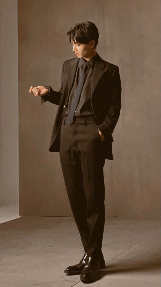 a man in a black suit stands in a room. he is looking down at the floor with a serious expression on his face. the walls of the room are a light brown. the light in the room is soft and warm. the man is dressed in a black suit with a white shirt and a dark tie. he is wearing black dress shoes. the suit is tailored to his body and fits perfectly. his stance is confident.