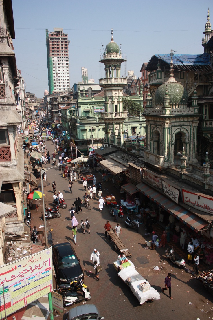 a highangle, long shot of a bustling street scene in mumbai, india.  the focal point is a beautiful green and white mosque situated amidst a dense collection of buildings, both old and new. a highrise building under construction is visible in the background. the street is filled with people, vehicles (cars, motorcycles, rickshaws), and street vendors. the overall atmosphere is one of vibrant activity and a blend of traditional and modern architecture. the image captures the energy and complexity of urban life in mumbai.