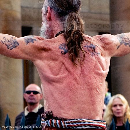 the image captures the back of a muscular, shirtless man with long hair tied back, likely a street performer. he has multiple tattoos on his back and arms and is facing away from the camera with his arms outstretched. he wears a patterned cloth wrapped around his waist. the background shows a blurred glimpse of a crowd watching him, suggesting an outdoor performance. the lighting suggests a sunny day, casting shadows across the scene.