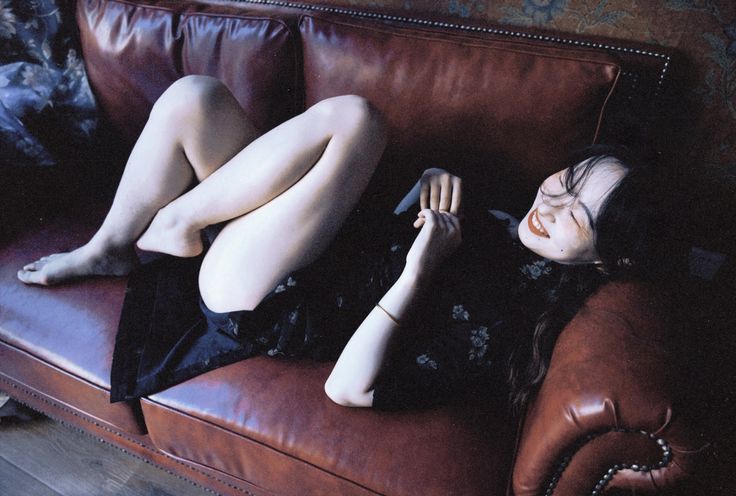 the image shows a young woman with long dark hair lying on a dark brown leather couch. she's wearing a darkcolored, possibly black, floral dress and is barefoot. her legs are crossed, and she appears relaxed and happy, smiling slightly. the lighting is somewhat dim, lending a sensual and intimate mood to the photograph. the style of the photograph and the setting evoke a sense of vintage or retro aesthetics, perhaps even hinting at art deco influences. the overall feel is candid and natural, capturing a moment of relaxed beauty.