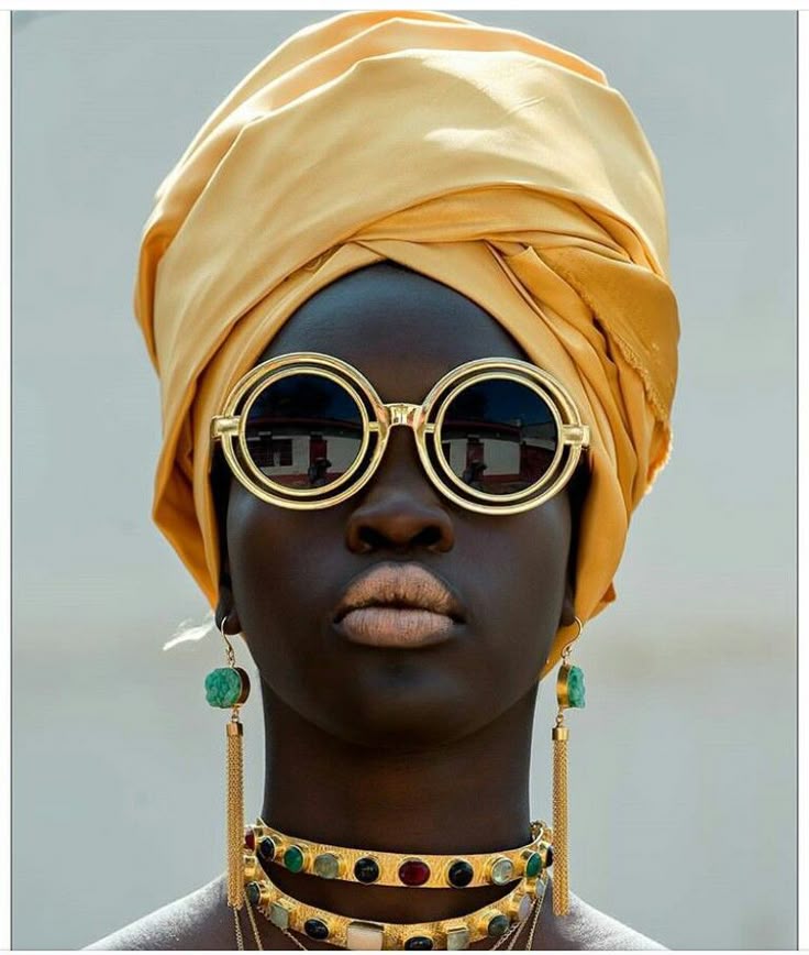 a portrait of a woman with dark skin and a yellow turban. she is wearing round sunglasses with gold frames, and gold jewelry including a layered necklace and dangling earrings. she is posing against a light gray background. the image is a studio shot.