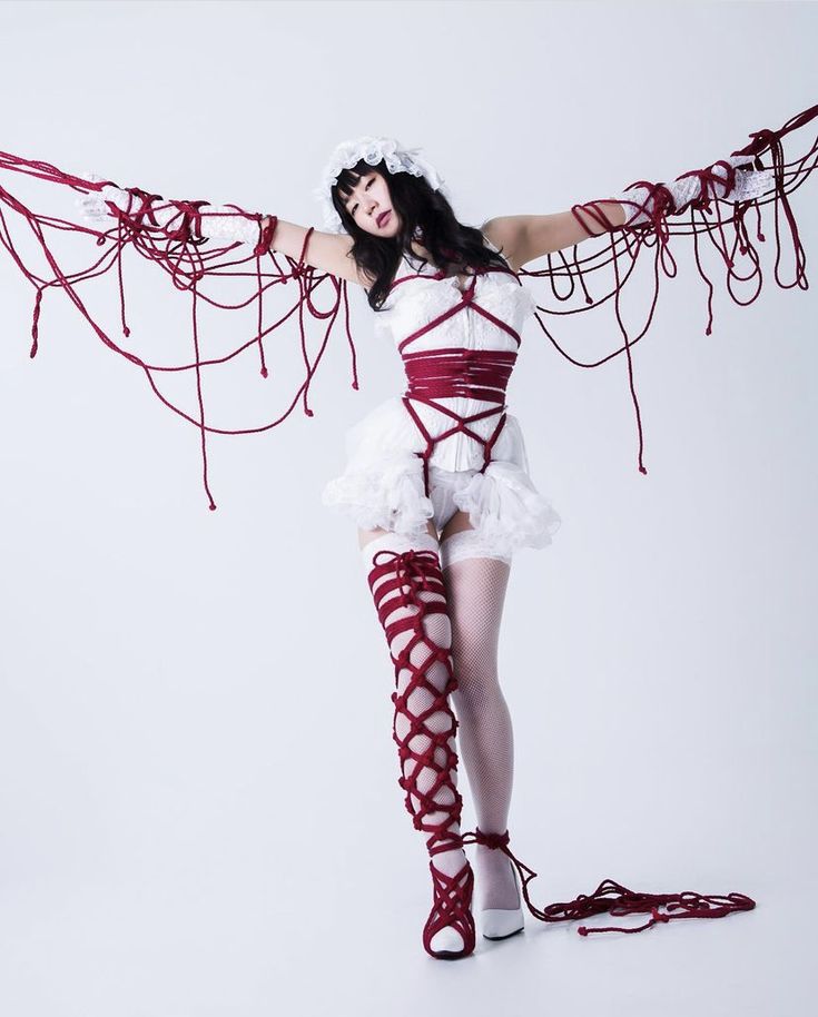 a full shot of a young woman, possibly a japanese model, posing in an intricate shibari rope bondage outfit.  she's wearing a white dress with a corsetstyle top, and her body and legs are adorned with elaborate red rope patterns. the ropes extend outwards from her body, creating a winglike effect. her makeup and hair are styled elegantly, complementing the artistic nature of the image. the overall aesthetic is a mix of fashion, art, and subtle sensuality, set against a plain white backdrop that emphasizes the subject. the photograph appears to be professionally taken in a studio setting.