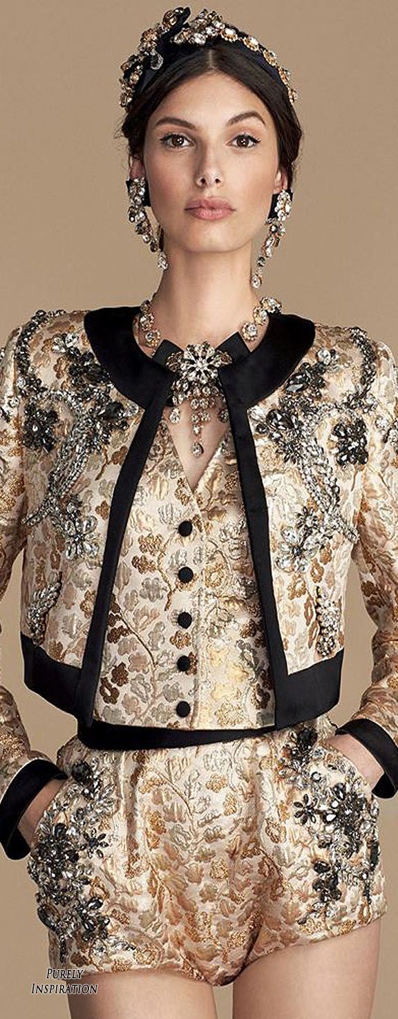model is posing in a studio shot wearing a dolce & gabbana outfit. the outfit consists of a gold brocade embellished shorts suit with a black trim jacket and matching accessories. the model wears a jeweled headband, earrings and necklace.  the background is a plain beige. the overall style is high fashion, glamorous and elegant.