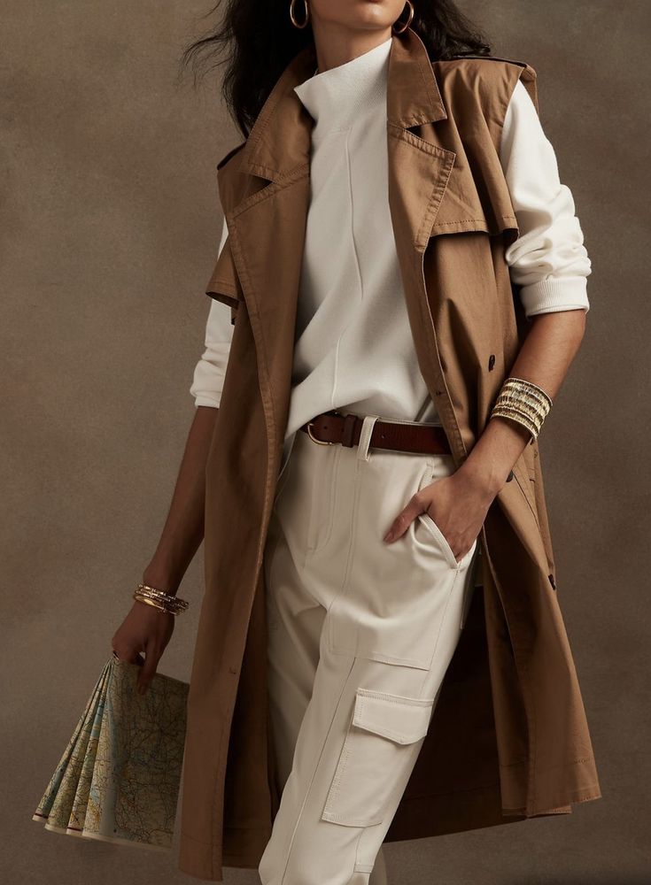 a fashion model is showcasing a stylish outfit. she is wearing a brown sleeveless trench coat over a white turtleneck sweater and offwhite cargo pants.  she accessorizes with a brown belt, gold bracelets, and gold earrings.  she's holding a folded map. the background is a muted brown tone. the overall style is casual yet chic and elegant.