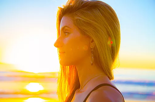 a blonde woman with a bright smile looks out at the golden sunset in the ocean. the image is a portrait and is taken close up and from her profile. the woman is wearing a black tank top and has long blonde hair with highlights. the background is a beautiful, golden sunset, which is reflected in the ocean water and the sky. the image is warm and inviting, with a feeling of happiness and peace.