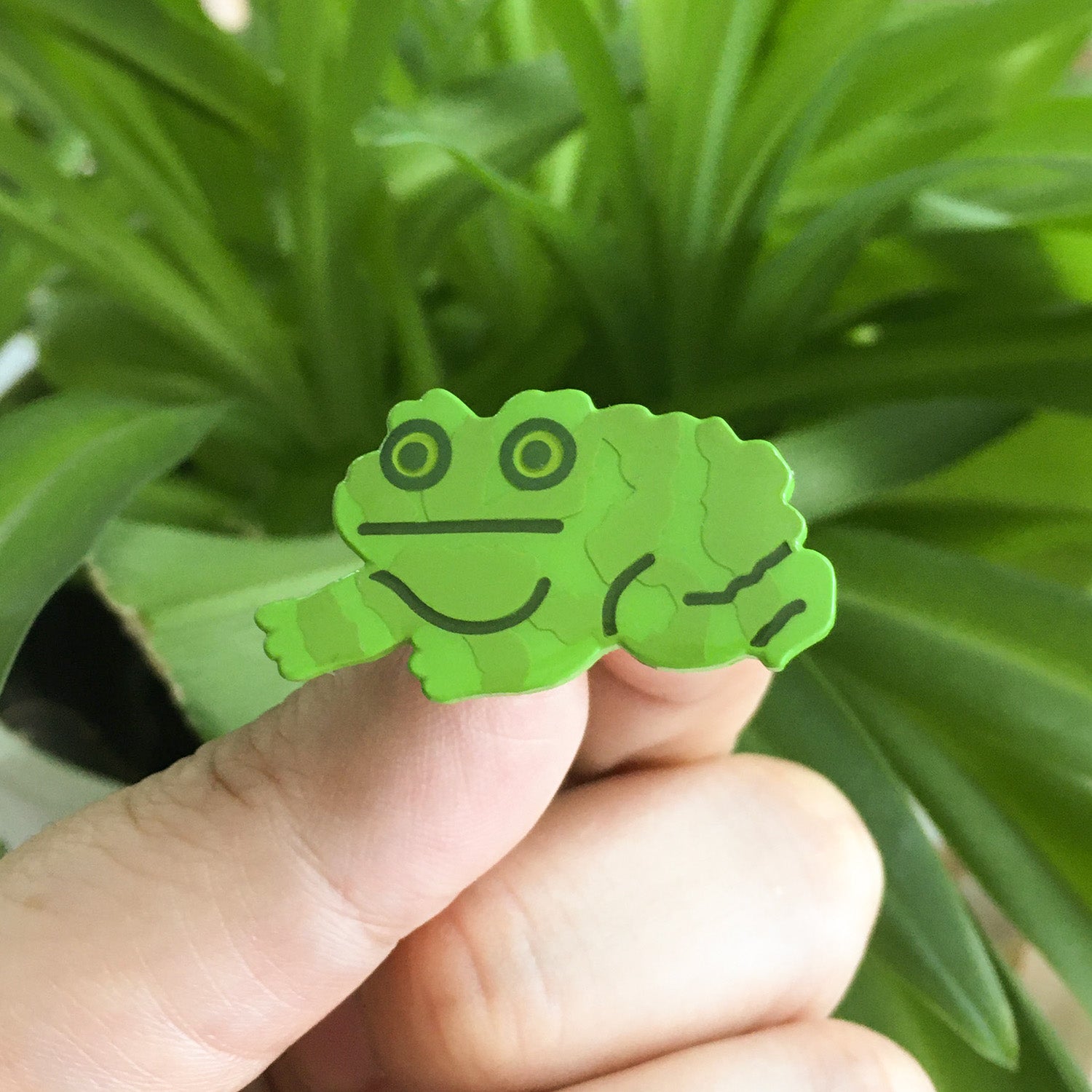 a closeup shot shows a person's hand holding a small, bright green enamel pin shaped like a cartoon frog. the frog has a textured, slightly bumpy surface resembling a toad's skin. it's simple in design with dark green outlines. the background is blurred but shows a lush green plant with long, spiky leaves, suggesting an outdoor or plantfilled indoor setting. the overall aesthetic is cute and whimsical.