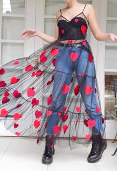 a young woman is wearing a fashionable outfit. the woman is standing in front of a white background, and she is wearing a black crop top with red hearts. she is also wearing denim jeans and a long black tulle skirt with red hearts. she is wearing black boots and has a trendy and beautiful look.