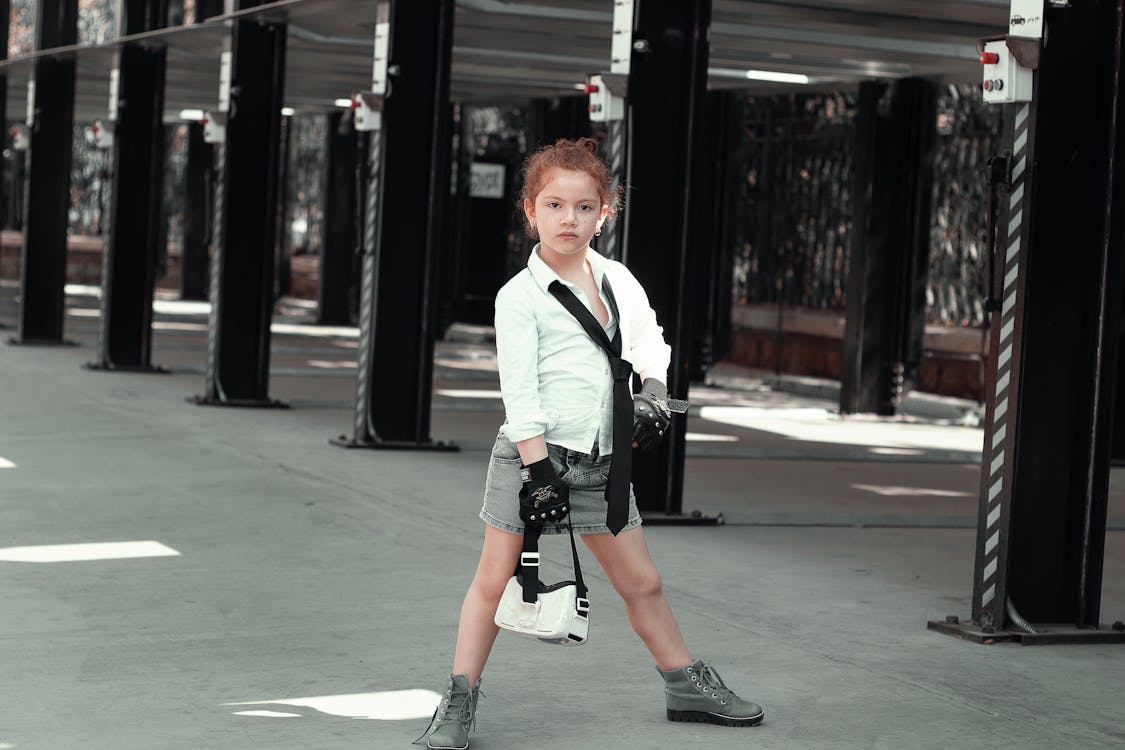 a young girl with red hair is striking a pose in a parking garage. she's dressed in a stylish outfit that includes a white shirt, black tie, denim skirt, gloves, and boots. she carries a small white bag and exudes a confident and cool attitude. the setting is urban and industrial, creating a contrast with her fashionable attire.