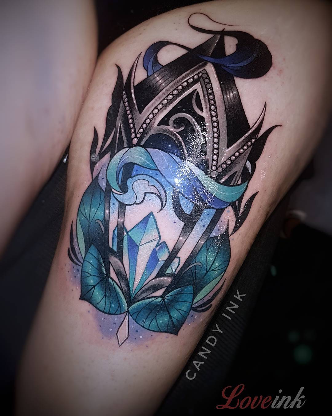 a person's thigh with a neotraditional style tattoo of a lantern with a crystal inside. the lantern has decorative metalwork and is surrounded by flames.  there are stylized leaves underneath the lantern.  the tattoo is done in shades of blue, teal, black and grey.  there is text next to the tattoo that says "candy ink" and "love ink".