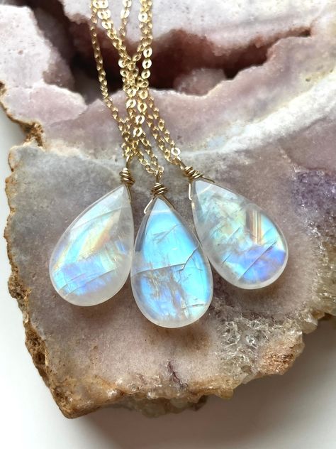 a closeup shot showcases three delicate gold necklaces, each featuring a shimmering, iridescent moonstone pendant in a teardrop shape. the stones exhibit a captivating play of light, reflecting hues of blue, white, and hints of peach. they rest against a backdrop of a raw, textured rock, likely quartz, highlighting the natural beauty of both the gemstones and their setting.