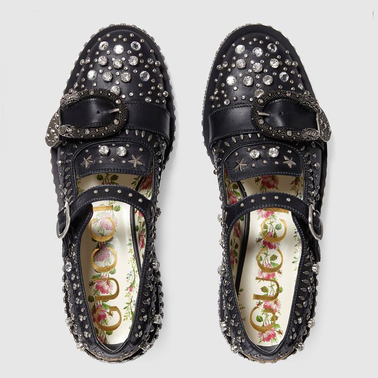 the image shows a pair of women's gucci shoes from a bird'seye view.  the shoes are black leather mary janes with a platform sole. they are heavily embellished with silvertoned studs, metallic stars, and clear crystals.  each shoe features a large ornate silver buckle.  the inside of the shoes shows a floral guccibranded insole.  the overall style is rockchic or punkinspired with a luxurious feel.