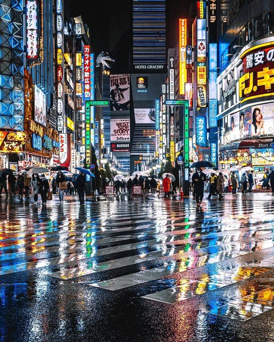 a vibrant, bustling street in tokyo at night. the street is wet with rain, and the neon lights of the surrounding buildings reflect colorfully on the ground. a crowd of people walk through the street, their umbrellas adding to the dynamic scene.