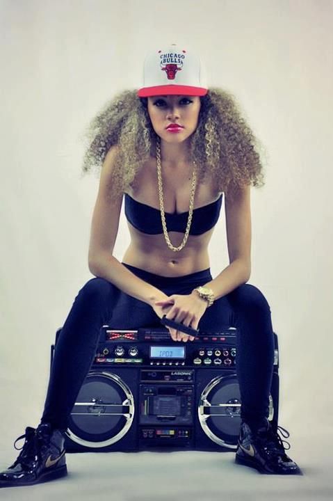 a woman is sitting on a large boombox,  wearing a black cap,  black pants,  black boots, and a large gold chain. she's posing in a studio setting.
