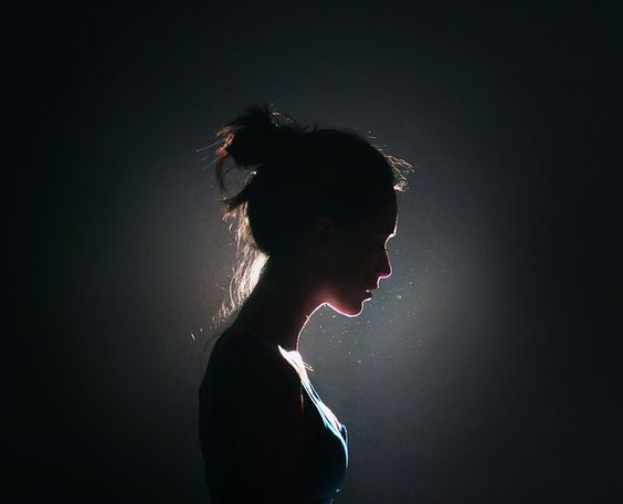 the image showcases the silhouette of a woman's profile set against a dimly lit background. the woman's hair is styled in a bun, and her head is slightly bowed. the lighting accentuates the contours of her face and creates a halo effect around her hair. the overall mood of the image is one of contemplation and introspection.