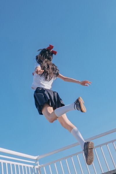 a girl in a school uniform is jumping in the air. she has a bow in her hair and is wearing a skirt, socks, and shoes. the sky is blue and there is a fence in the background. it is a sunny day.