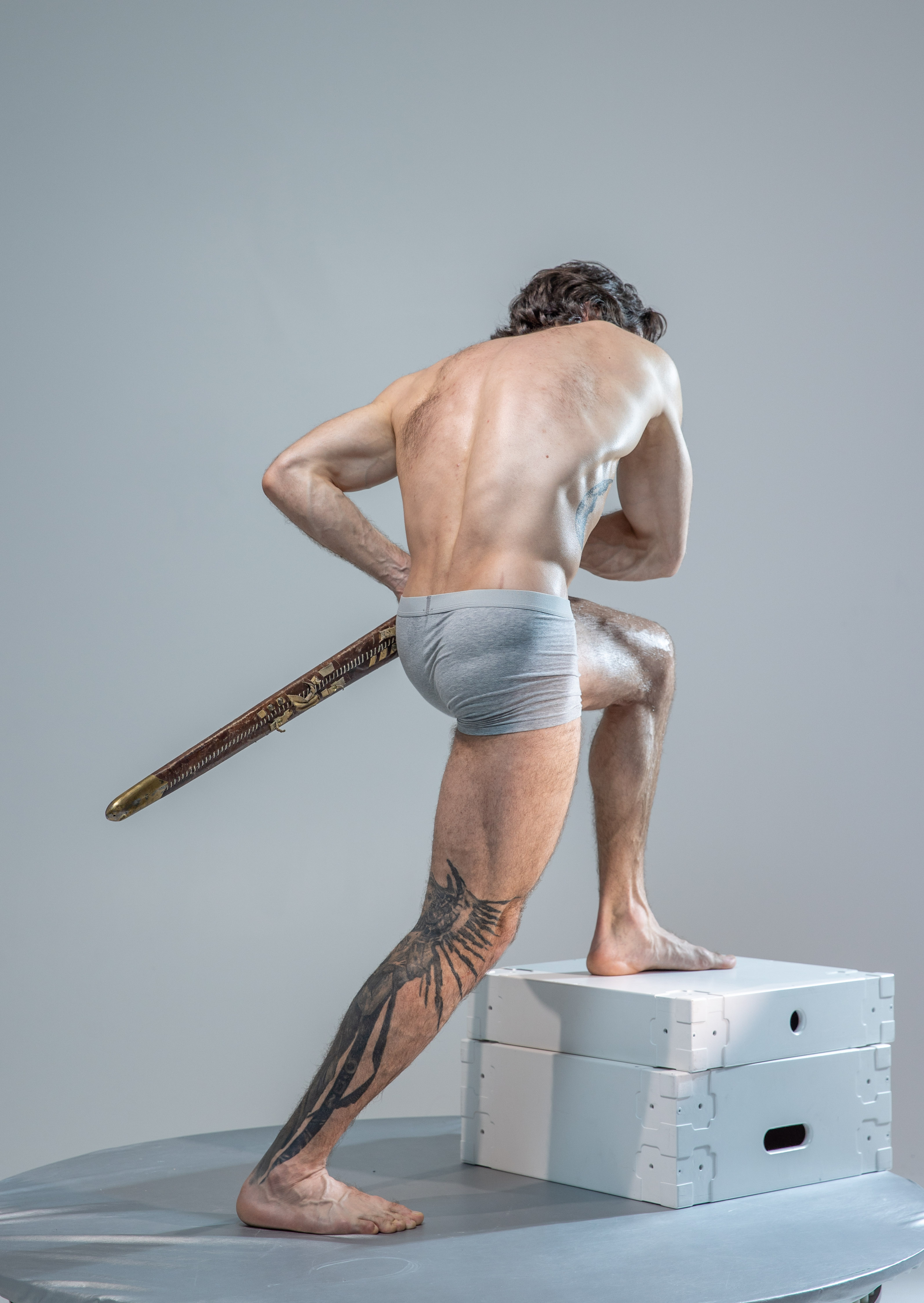 a muscular man with flowing hair stands in a dynamic pose on a white platform, his back to the camera. he holds a sword in a defensive stance, its blade angled downwards. his body is illuminated by soft studio lighting, highlighting his defined musculature and the tattoo on his leg. he wears only grey underwear, allowing for a clear view of his physique. the neutral background and minimal props draw focus to the man's form and the energy of his pose.