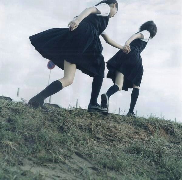 two girls in school uniforms are holding hands and running down a grassy hill. they are wearing black dresses, white shirts, black socks, and black shoes. the sky is overcast.