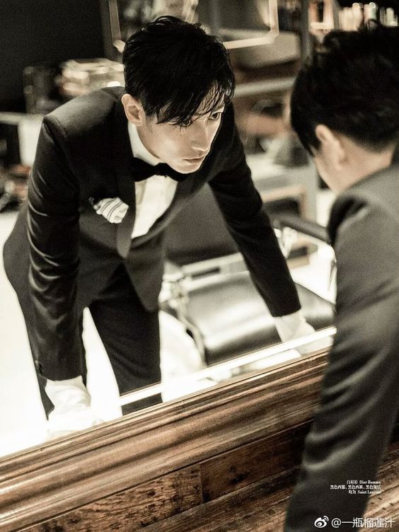 a man in a tuxedo is looking intensely at his reflection in a mirror. he is leaning forward, with his hands on the frame of the mirror. the photo appears to be in black and white.