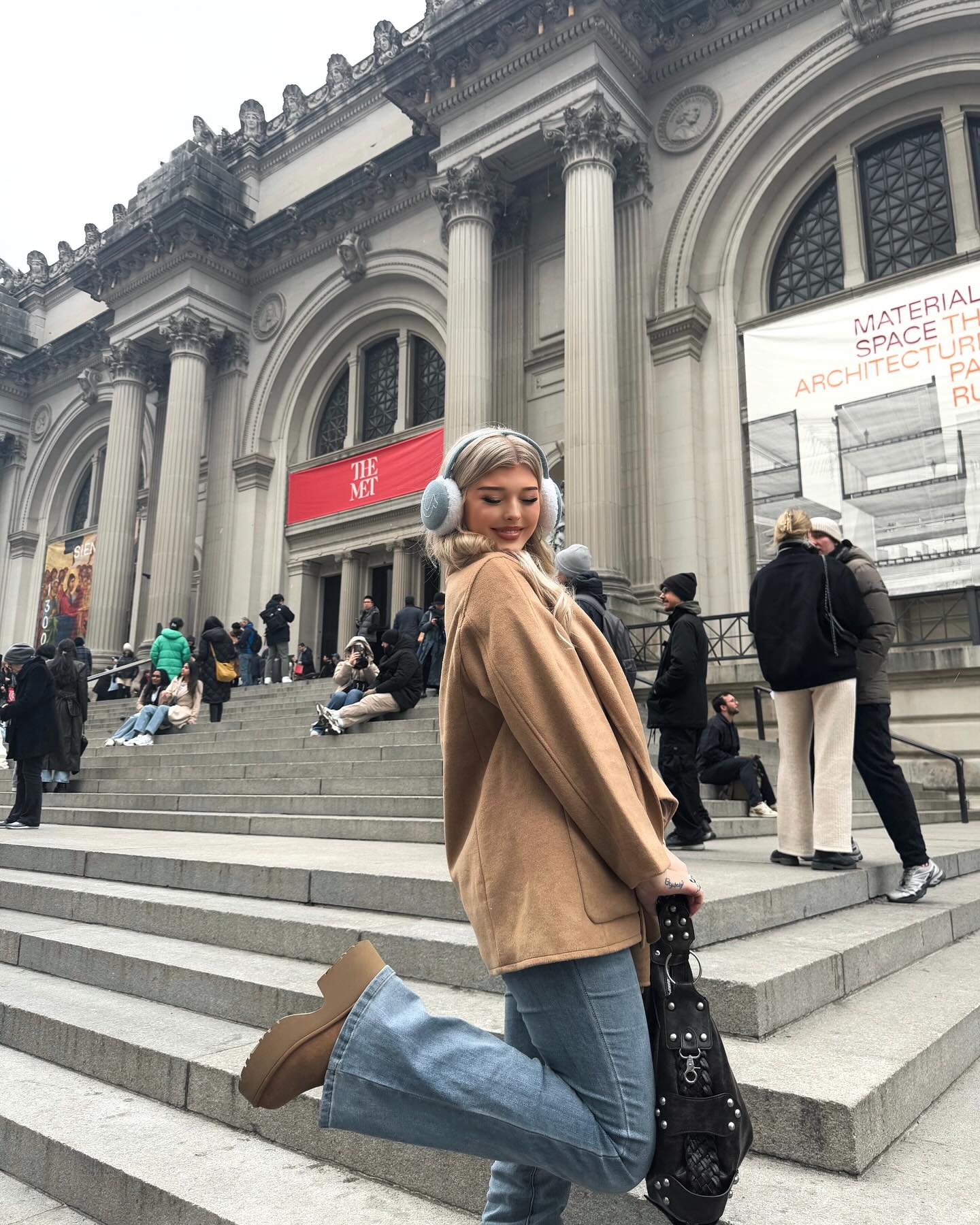 a young woman posing on the steps of the metropolitan museum of art. she is wearing earmuffs, a brown coat, blue jeans, and brown platform boots. she is also carrying a studded black handbag. people are sitting on the steps behind her.