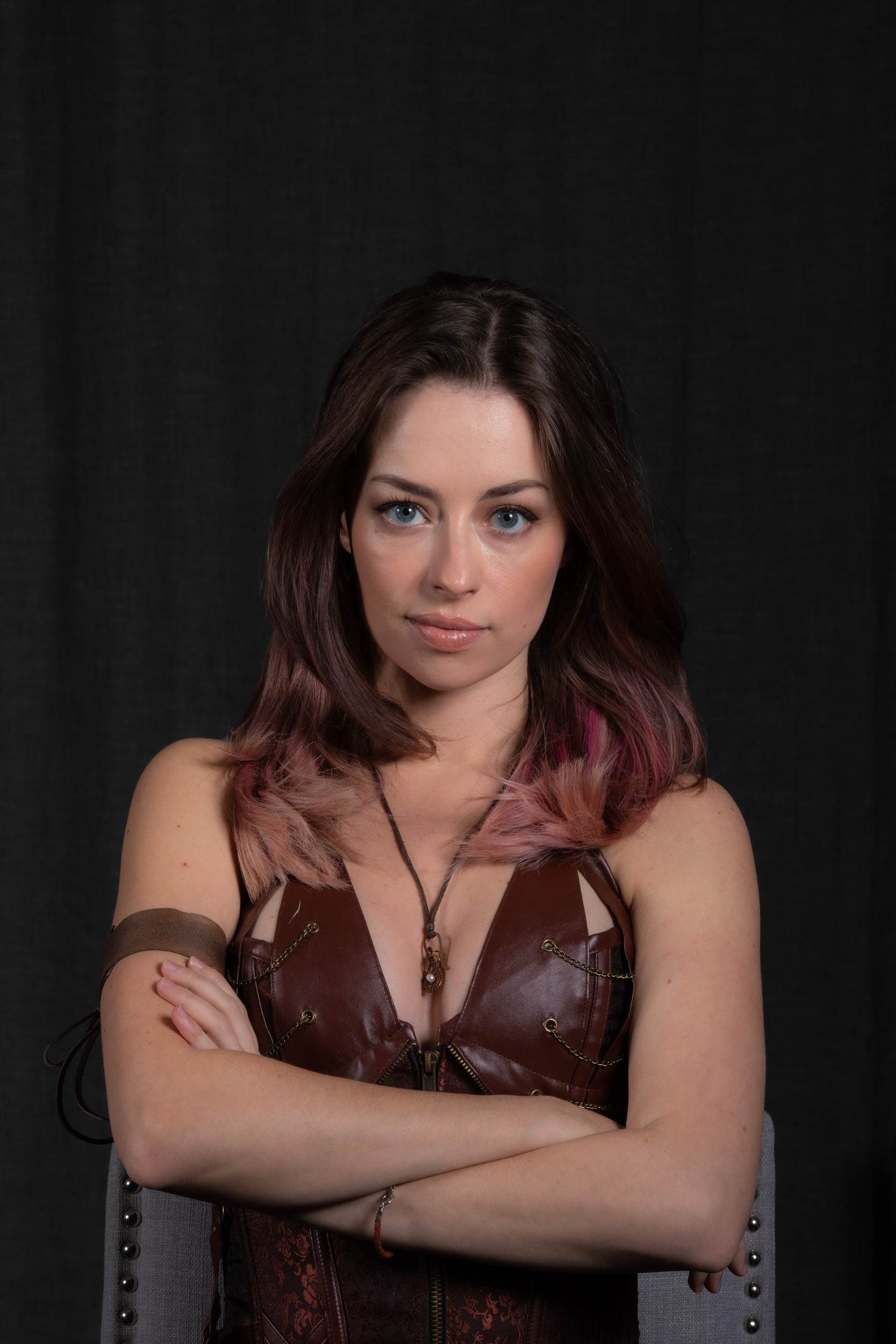 a young woman with pinktinged brown hair confidently poses for a portrait against a black backdrop. she wears a brown leather steampunkinspired corset and a matching arm cuff. her arms are crossed, and she gazes directly at the camera with a neutral expression. the image evokes a sense of strength and individuality.