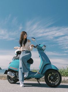a woman in jeans and a white top sits on a teal vespa scooter, facing the camera. she's parked on a paved road near a beach, with the blue ocean and sky visible in the background. the sky is filled with wispy white clouds and the image is brightly lit by natural daylight.