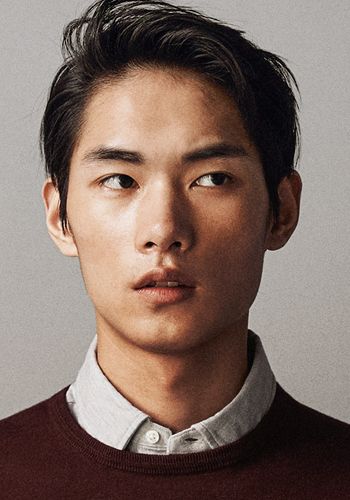 a portrait of an asian man wearing a maroon sweater over a light gray buttondown shirt. he has dark hair styled with a slight wave, and he is looking slightly off to the side with a serious expression. the background is a neutral, light gray color.
