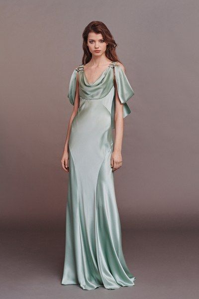 a fulllength studio shot of a model wearing a pale green, silk maxi dress.  the dress features a cowl neck, sleeveless design with delicate shoulder straps, and a flowing, elegant silhouette. the model has long brown hair and a neutral makeup look. the background is a muted taupe or grey.