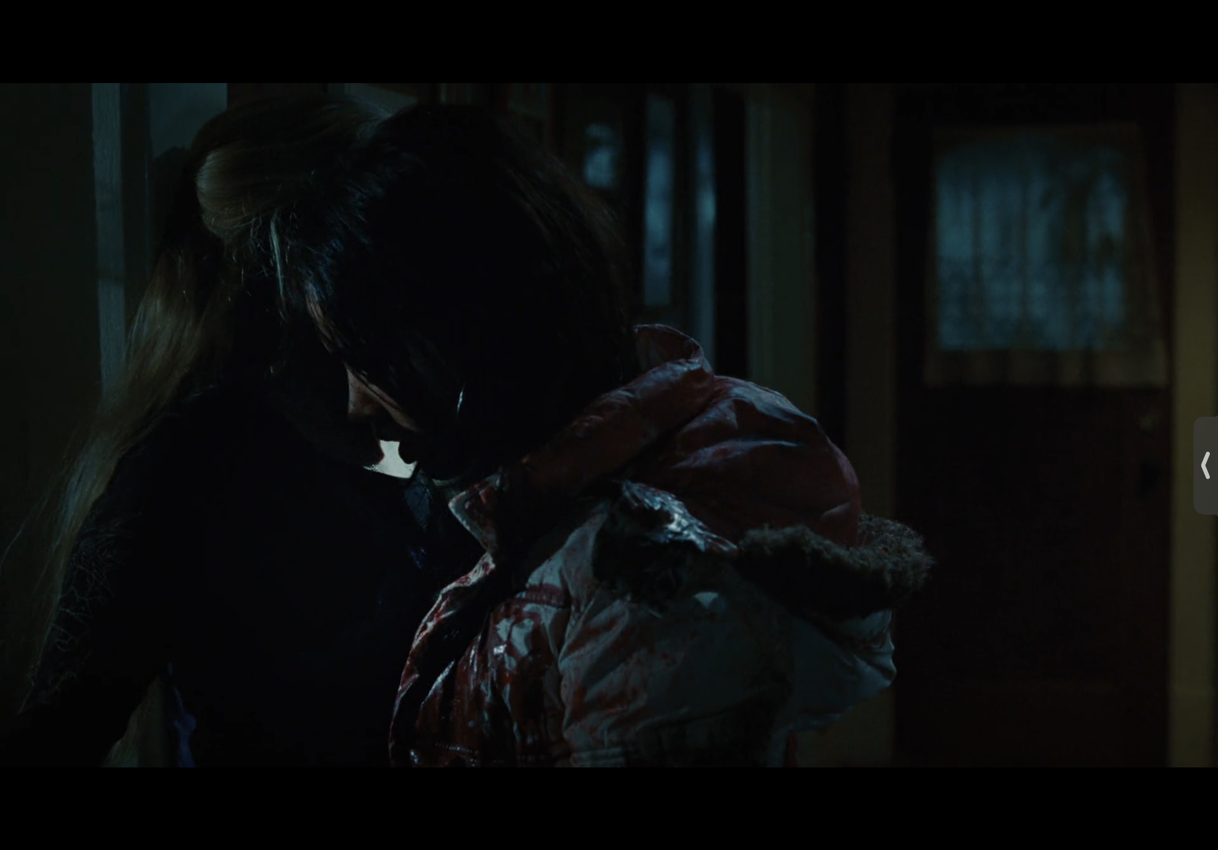 a closeup shot of a man and woman in a dark, dimly lit room. the man is covered in blood and appears to be injured or dead. the woman is standing close to him, her face is obscured by the shadows. the atmosphere is tense and sinister, suggesting a violent or horrific event has occurred. the scene likely comes from a horror or thriller film.