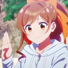 he image is a portrait of a young woman with long brown hair tied up with an orange ribbon. she is wearing a light blue hoodie and has bright yellow eyes. she is smiling and waving at the viewer with her left hand. the background is a blur of warm colors, suggesting a bright outdoor setting. the overall impression is one of happiness and friendliness.