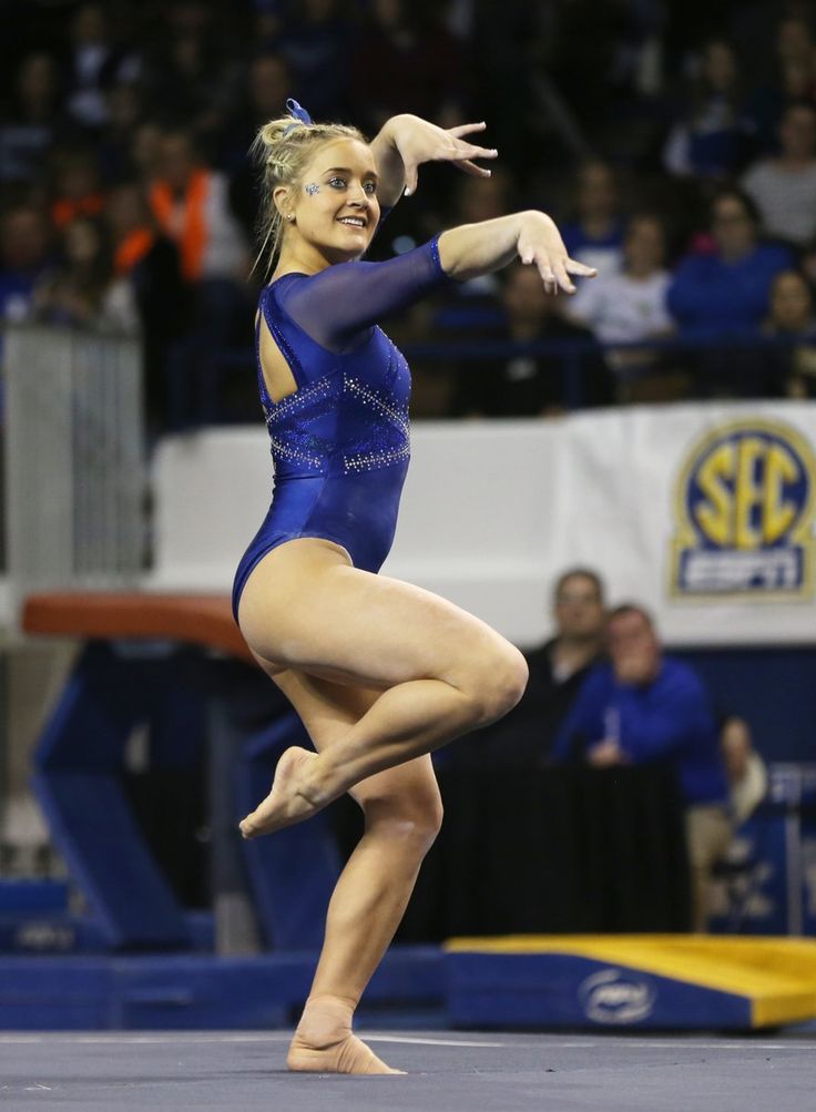 a female gymnast is performing a floor exercise in a gymnastics competition. she is wearing a blue leotard and has a confident expression on her face. she is midair, completing a trick with her body contorted in a complex pose, demonstrating incredible flexibility and athleticism.  the blurred background is the audience watching the competition.