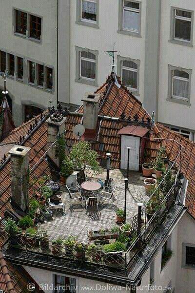 an overhead shot of a charming rooftop garden nestled atop an apartment building in a european city. the rooftop features a small patio with a wooden deck, a table and chairs, and numerous potted plants, creating a vibrant and serene outdoor space. the building itself has a red tile roof and is characteristic of european architecture.  the surrounding cityscape is visible in the background. the scene offers a glimpse into the creative use of limited space for gardening in an urban setting.