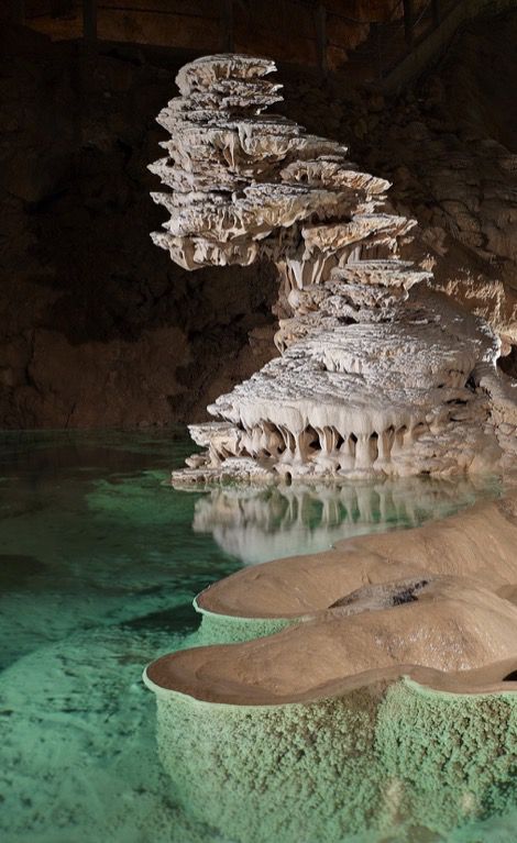 a large, intricate rock formation resembling a mushroom, covered in stalactites and stalagmites, sits in a cave filled with clear green water. the cave walls are dark and rough, contrasting with the smooth, white rock formations. the water reflects the light, creating a tranquil and ethereal atmosphere.