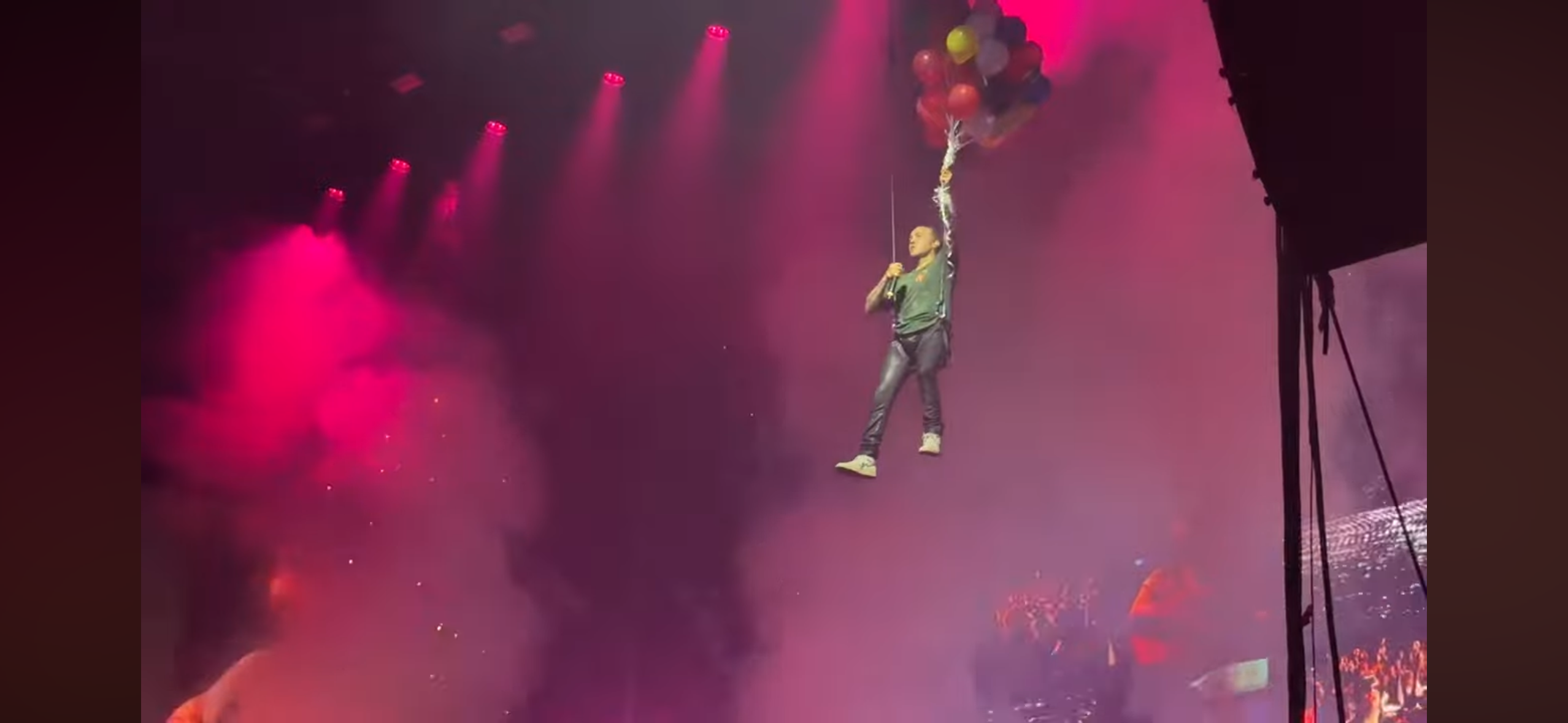 a man is suspended above a concert audience on a harness, surrounded by balloons. the stage is lit with pink and purple lights, and the crowd is visible below. he is wearing a dark green shirt and dark pants.