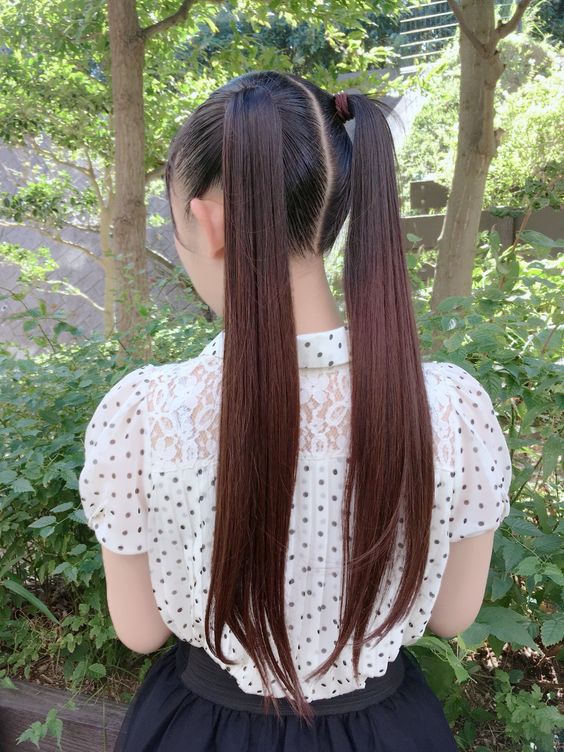 the image shows a woman from behind, standing outdoors in a parklike setting. she has long brown hair styled in two pigtails, and is wearing a white polka dot blouse and a black skirt. the background is filled with lush greenery, and the lighting is natural and bright.