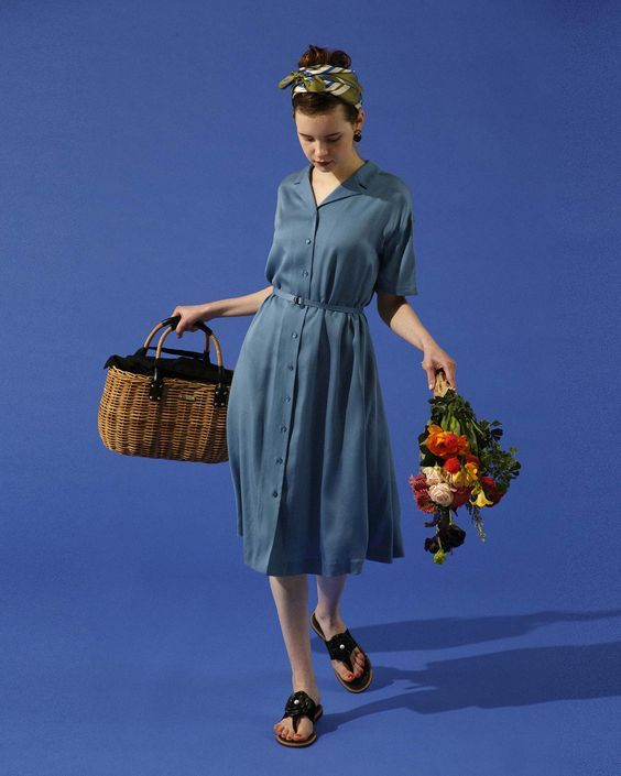a woman is wearing a blue dress, a blue headscarf, and black sandals. she is carrying a wicker basket with a black handle and a bouquet of flowers. she is standing in front of a blue background.