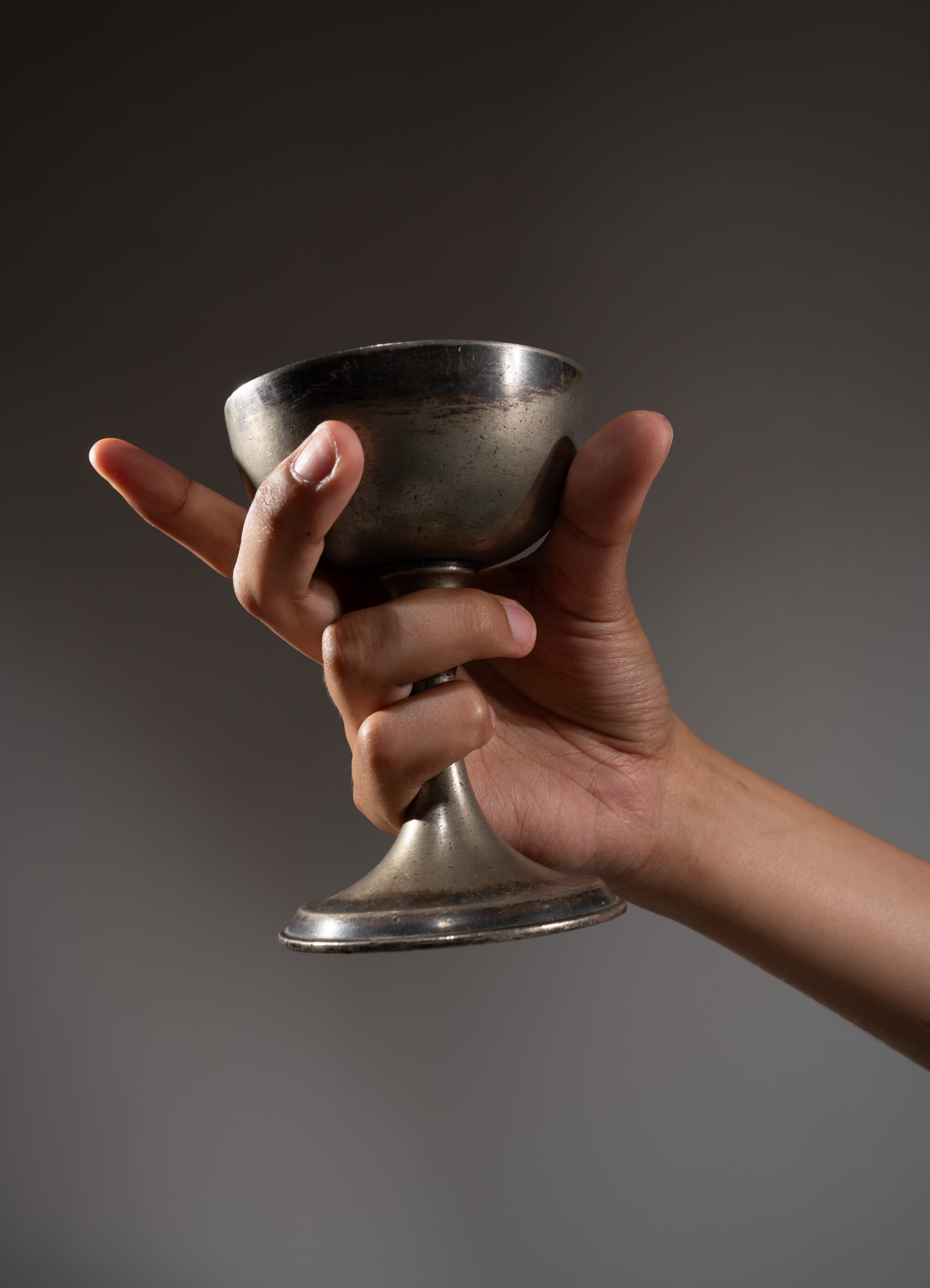 an adult hand holds a tarnished silver chalice against a gray background. the chalice has a simple, elegant design with a wide bowl, a slender stem, and a round foot. the lighting is soft and highlights the chalice's intricate details and tarnished finish.