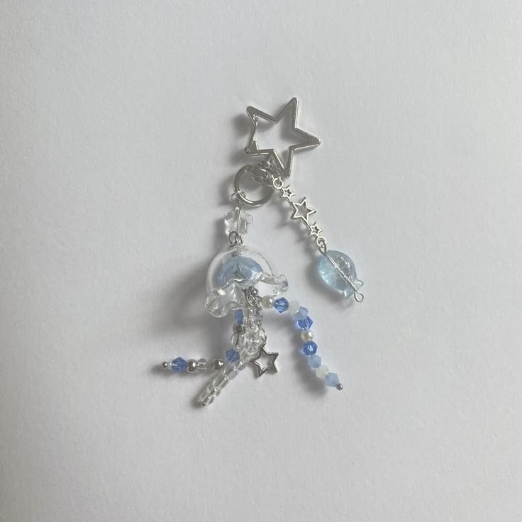 a handmade jellyfish charm keychain lies on a white surface. the charm features a clear jellyfishshaped dome with blue accents, adorned with strands of clear and blue beads mimicking tentacles. a silver star charm dangles among the beads. attached to the jellyfish is a silver ring connecting to a chain of small stars and a blue heartshaped charm, topped with a large silver star charm.