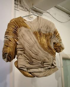 a closeup shot of a beige and gold dress, with the fabric forming pleats and layers, draped in a delicate and flowing fashion. the dress is designed with an intricate artistry and craftsmanship, giving it a unique and abstract appearance. the light hitting the fabric creates a beautiful play of shadows and highlights.  it appears to be a handmade garment.