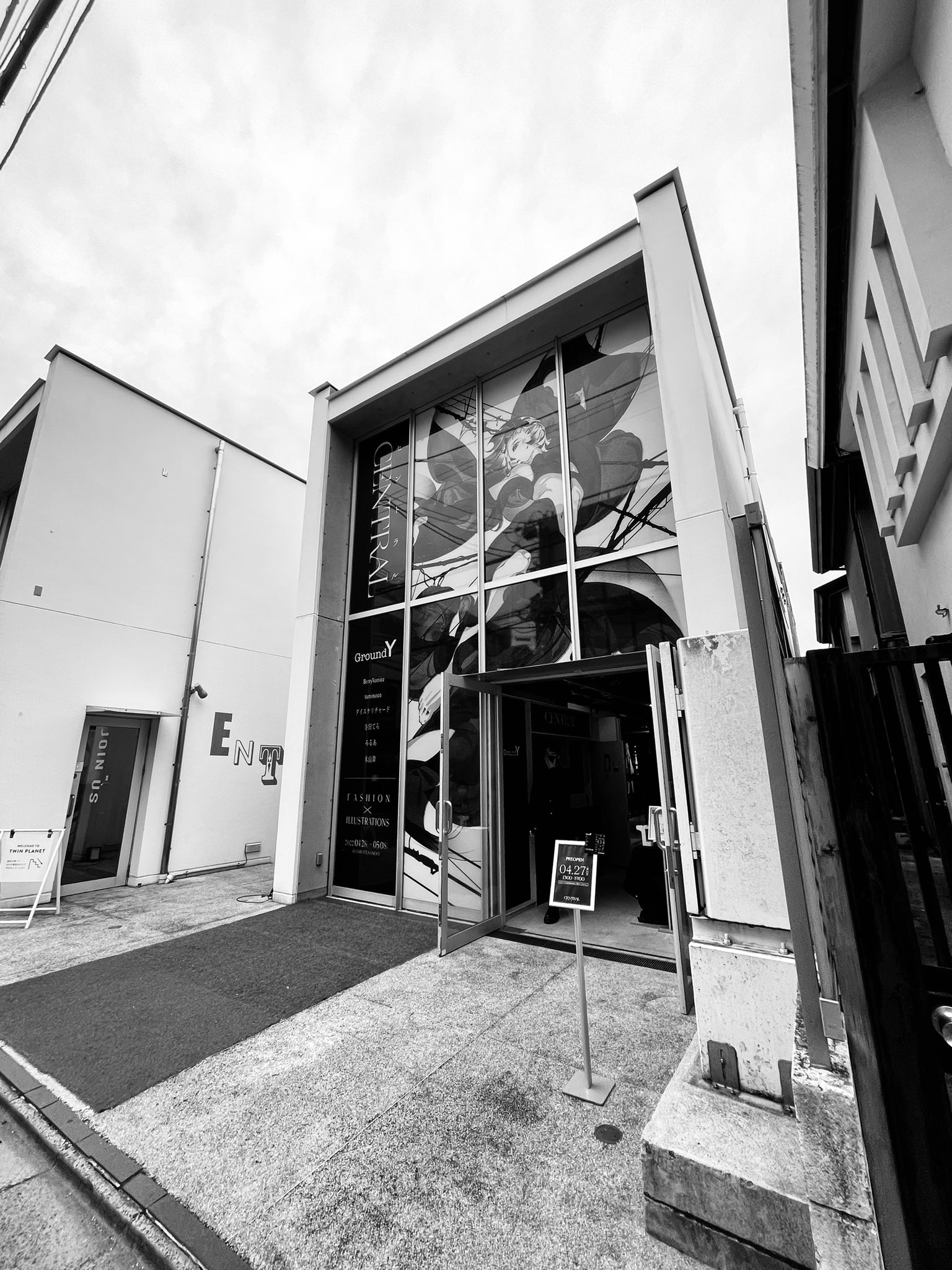 a black and white photo of the exterior of a modern art gallery. the building is white with large windows and a large poster of a woman on the windows.  the door is open and there is a person inside. the photo is taken from a low angle.