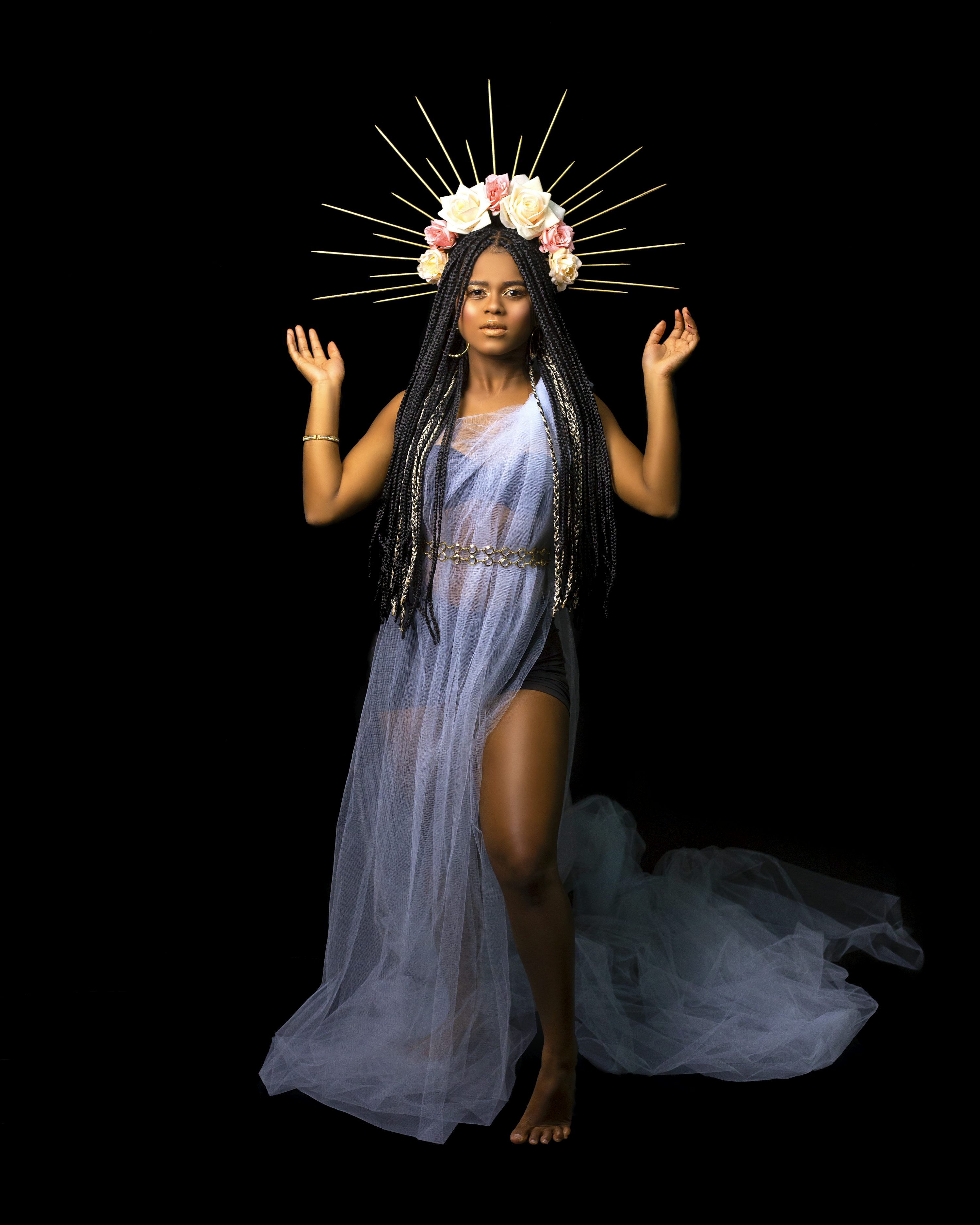 the image portrays a young black woman dressed in an ethereal, flowing lightblue tulle gown.  she wears a crown of white and pink flowers adorned with gold accents. her long black braids frame her face, and she stands with a graceful posture, arms raised slightly. the background is entirely black, making the woman and her clothing the focal point. the overall aesthetic suggests a mystical or goddesslike persona. the lighting is soft and accentuates the delicate texture of her dress and the details of her headpiece.